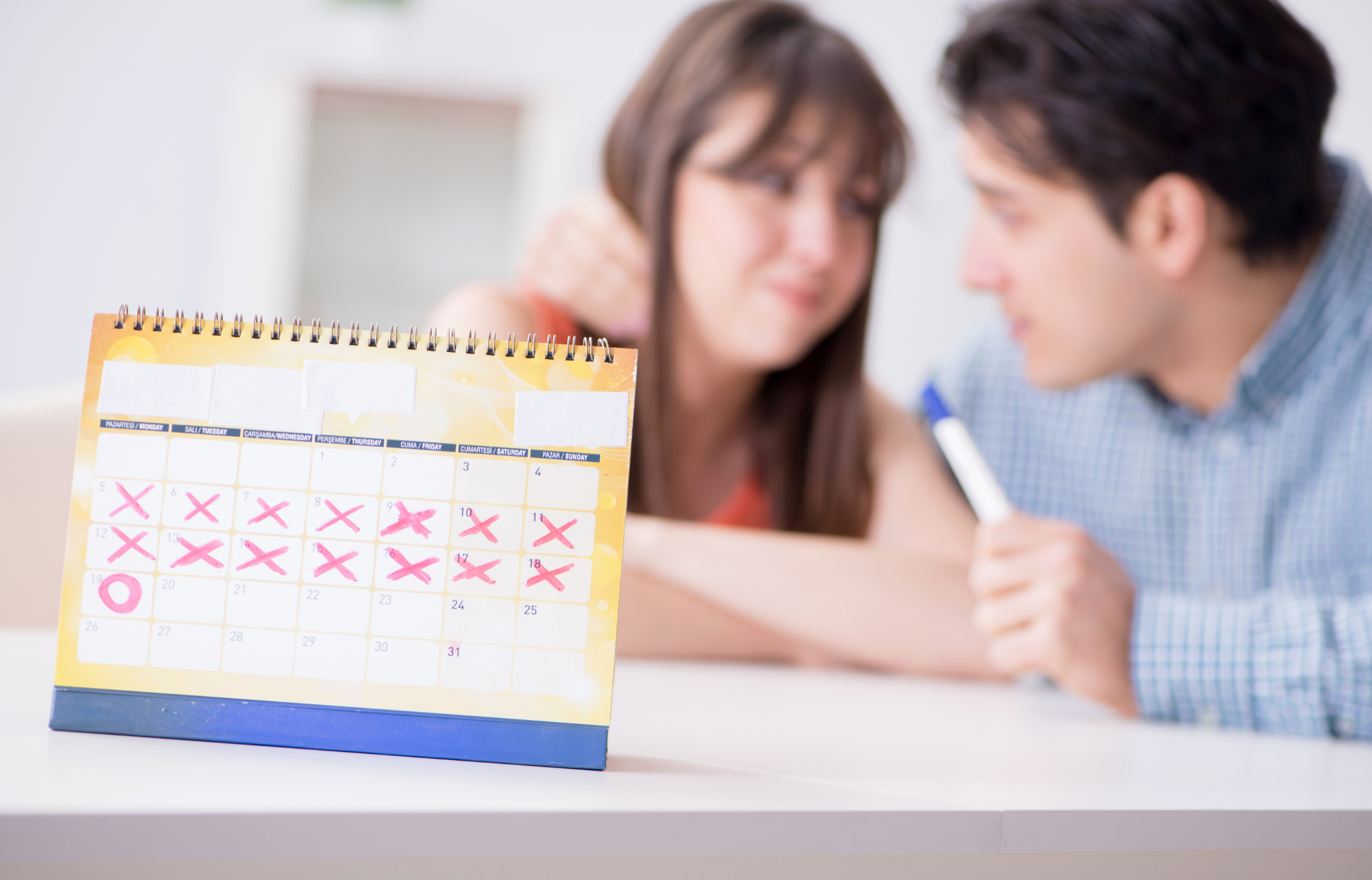 Kalendarz z zaznaczonymi dniami cyklu i para planująca ciążę w tle.